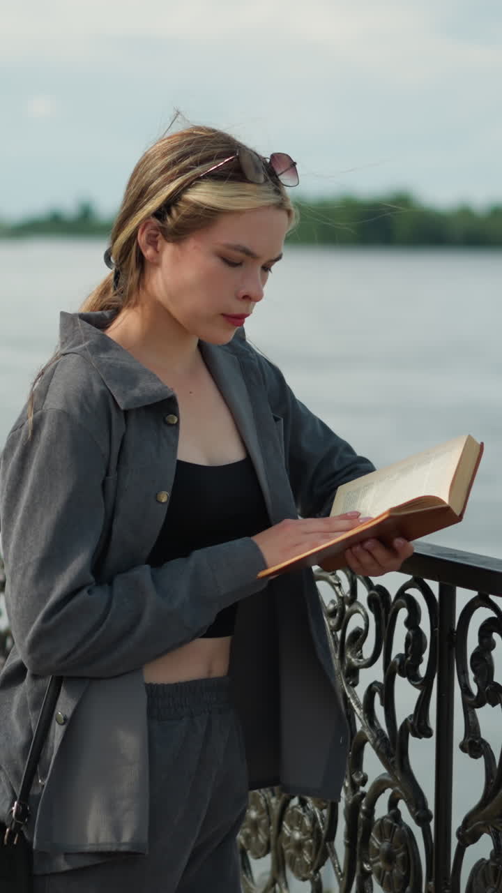 joven con ropa gris descansa su mano derecha en una valla de hierro mientras lee un libro, el viento sopla suavemente su cabello y ropa mientras lee, con árboles y la orilla del río visibles en la distancia