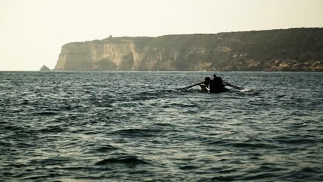 Silhouette-of-rowing-team-on-a-boat-in-front-of-a-sea-cliff-landscape-at-sunset