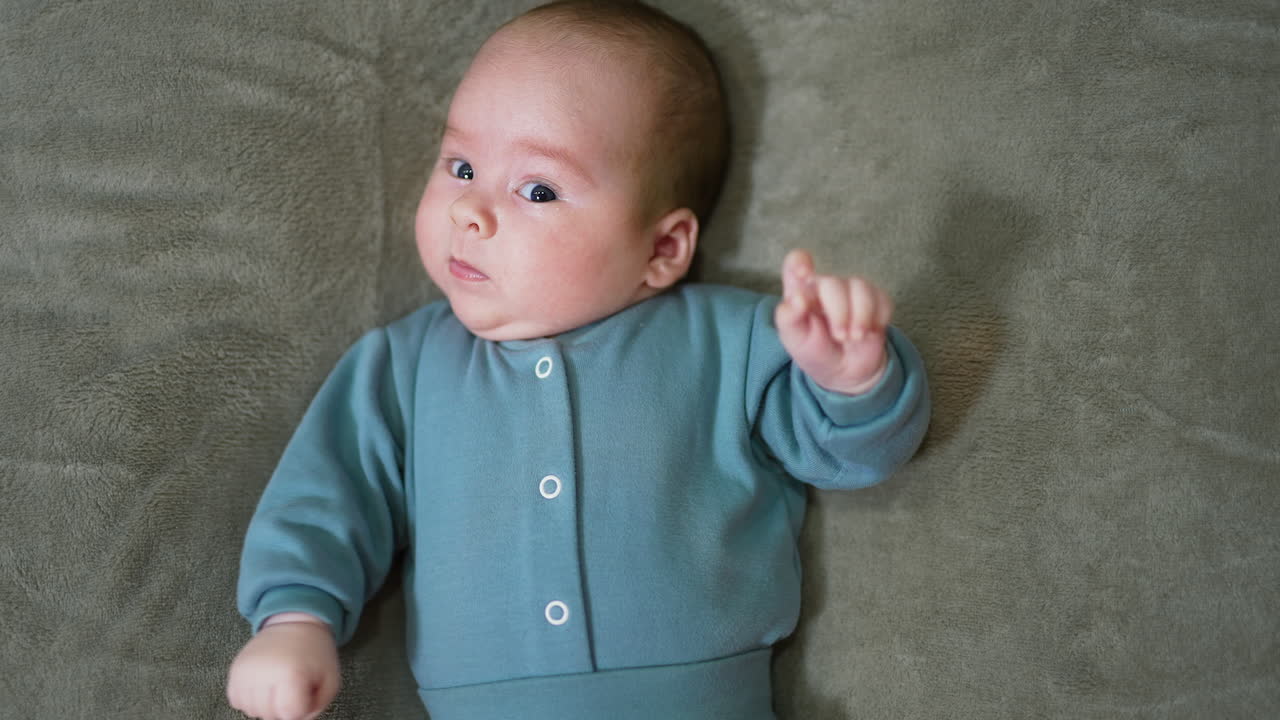 Adorable baby looking suspiciously at the camera. Sweet infant boy in blue clothes lying on the bed. Close up. Grey backdrop.