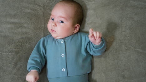 adorable baby looking suspiciously at the camera. sweet infant boy in blue clothes lying on the bed. close up. grey backdrop.
