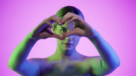 medium static shot of a pretty young woman folding her hands to a heart symbolizing love and mindfulness in front of pink background in slow motion