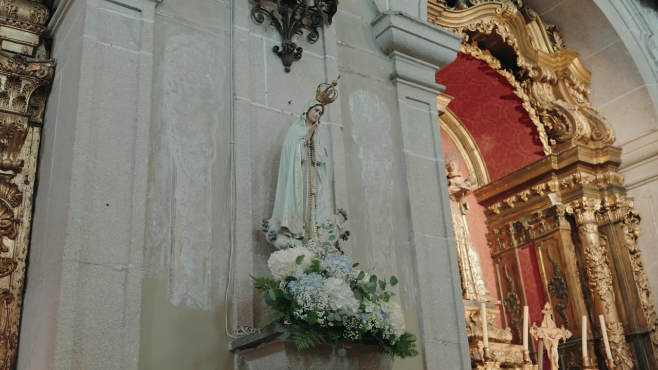 Estátua de Nossa Senhora de Fátima em uma parede de igreja com flores brancas ao lado de um altar barroco dourado