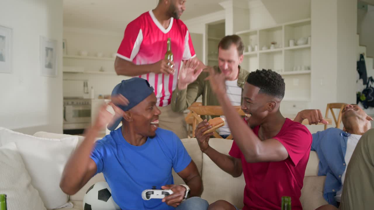 Amigos homens diversos pegando cachorros-quentes de uma cesta, jogando com controles durante uma partida de futebol.