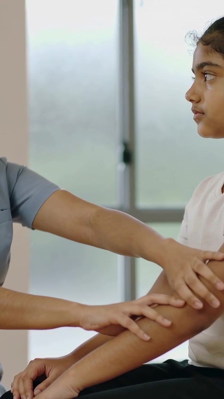 Video verticale: Infermiera in divisa medica che si protende e palpa il braccio di un adolescente su una sedia da visita per un esame, con un fermo della finestra.