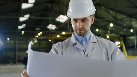 caucasian engineer wearing a helmet holding blueprint and looking around in a factory