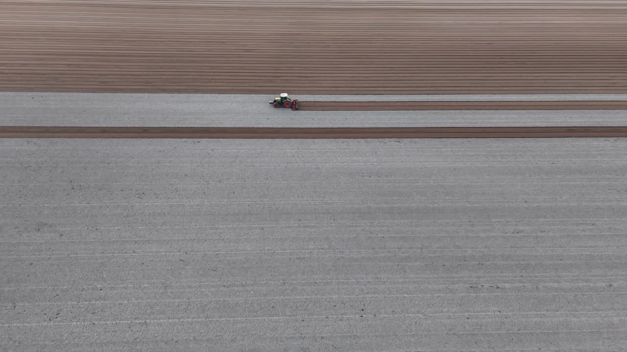 Drone’s minimal canvas captures lone tractor pulling a harrow in slow motion, drawing deep brown seed lines beside bleached soil furrows across an expansive field as warm dusk light fades into evening