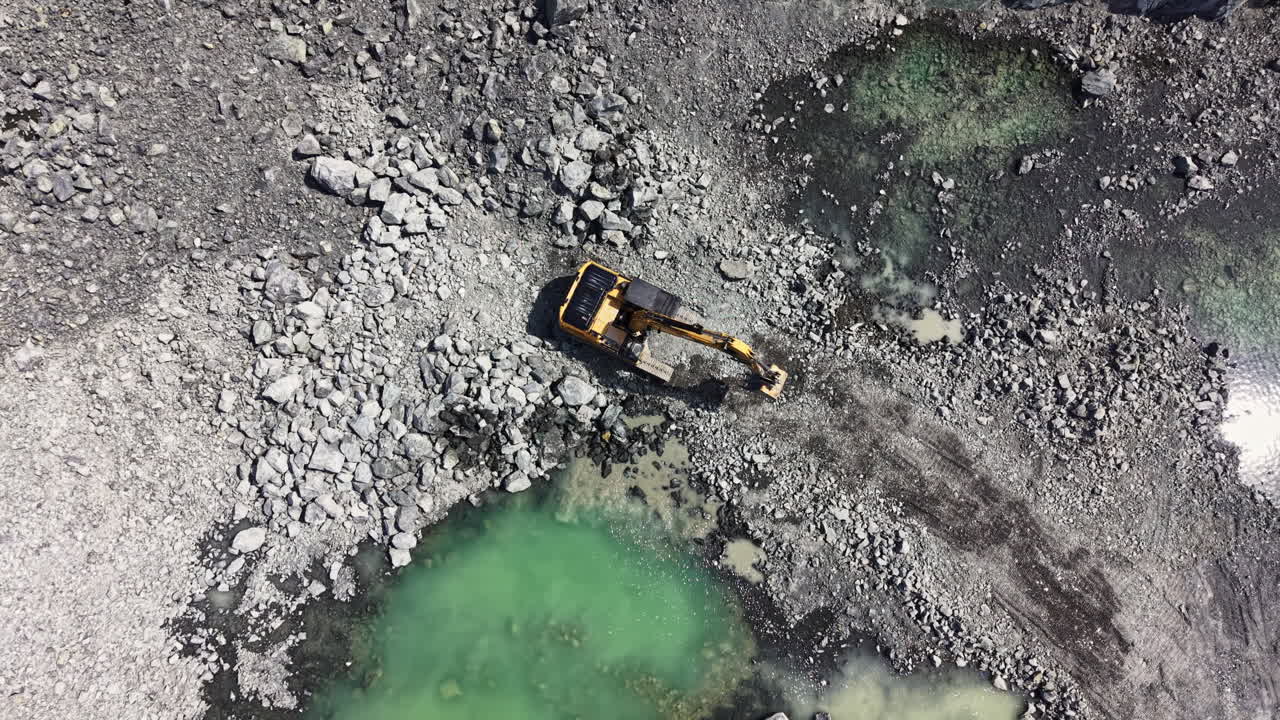 На гранитном карьере днем работает техника среди луж воды и обломков камней.