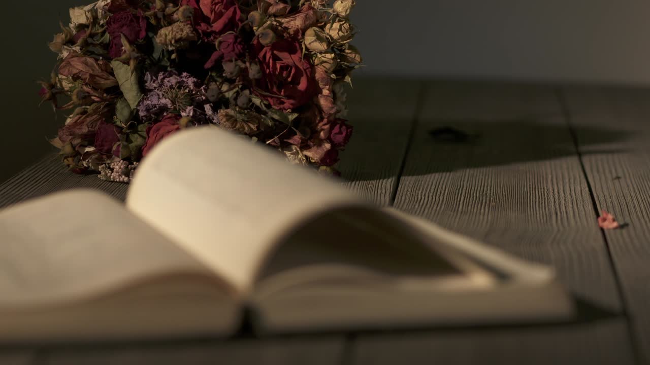 Bukett med visne blomster og åpen bok på et trebord i et mørkt rom
