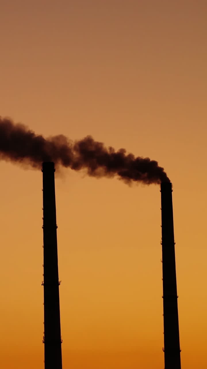 Dirty emissions on the orange sky background. Two pipes with dark smoke at sunset. Ecological disaster. Air pollution. Vertical video