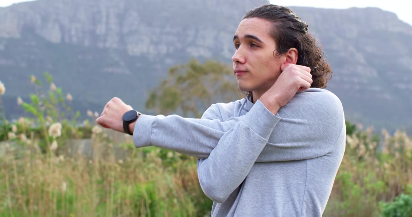 Uomo che fa stretching incrociando le braccia, riscaldandosi per un'escursione in un prato, con felpa grigia con cappuccio e smartwatch