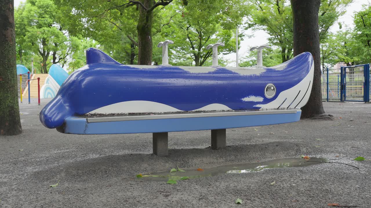 A side-view shot of a blue whale-shaped spring rider on a wet, gravel playground in an urban park