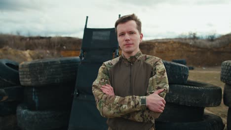retrato de un militar confiado en un uniforme verde camuflado que dobla los brazos en su pecho y posa con una expresión seria cerca de barricadas hechas de neumáticos de automóviles en la estepa durante el entrenamiento de combate