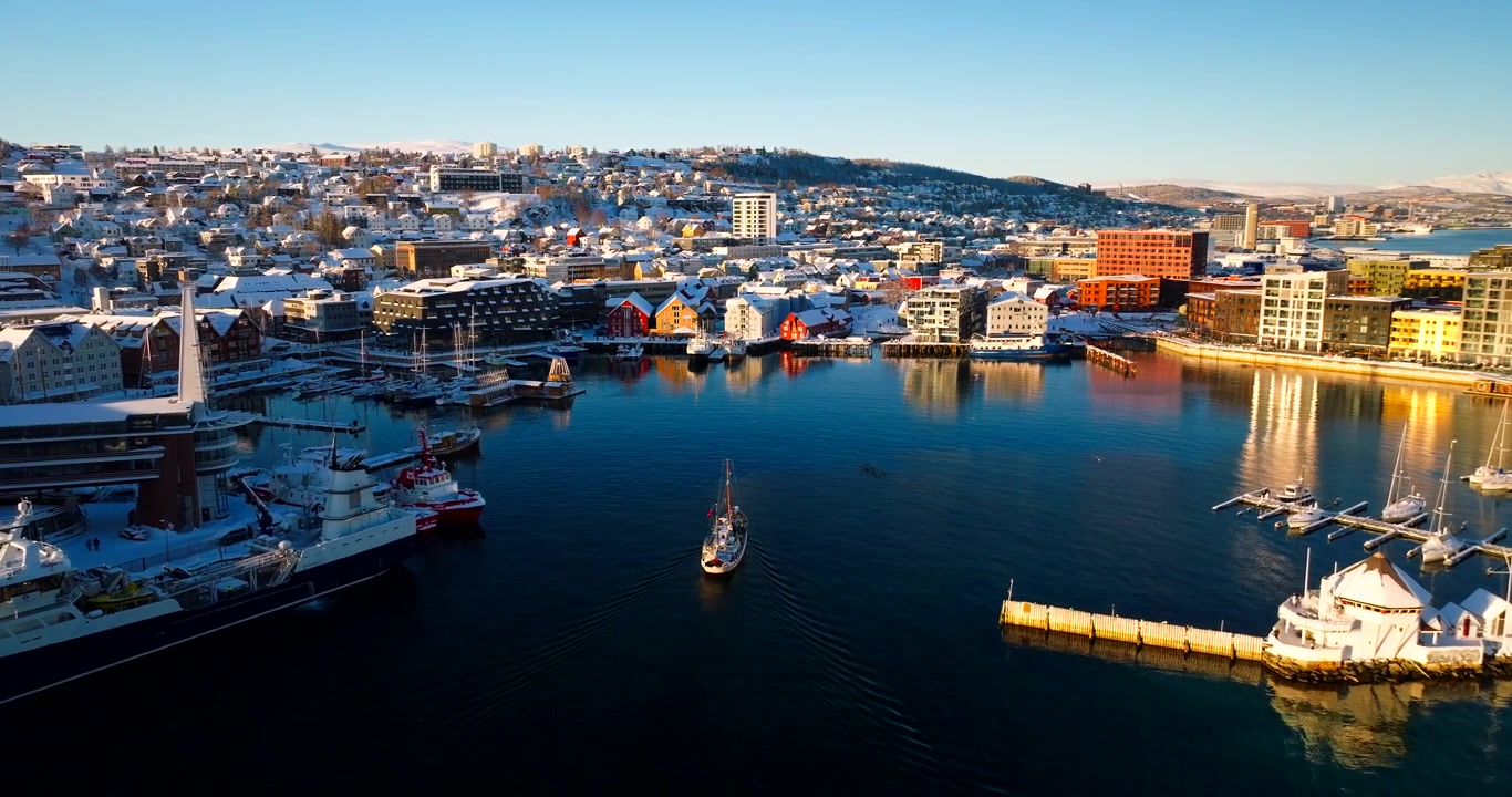 Drohnenansicht des historischen Bootes Hermes II, das in der Dämmerung in einen verschneiten Hafen in Tromsø, Norwegen, segelt. Vorwärtsflug aus der Luft