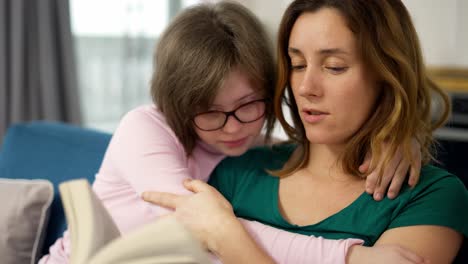 Junge-Mutter-Liest-Ihrer-Tochter-Mit-Down-Syndrom-Ein-Buch-Vor,-Während-Sie-Auf-Dem-Sofa-Im-Zimmer-Sitzt