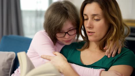 madre joven leyendo un libro a su hija con síndrome de down mientras están sentados en el sofá en la habitación