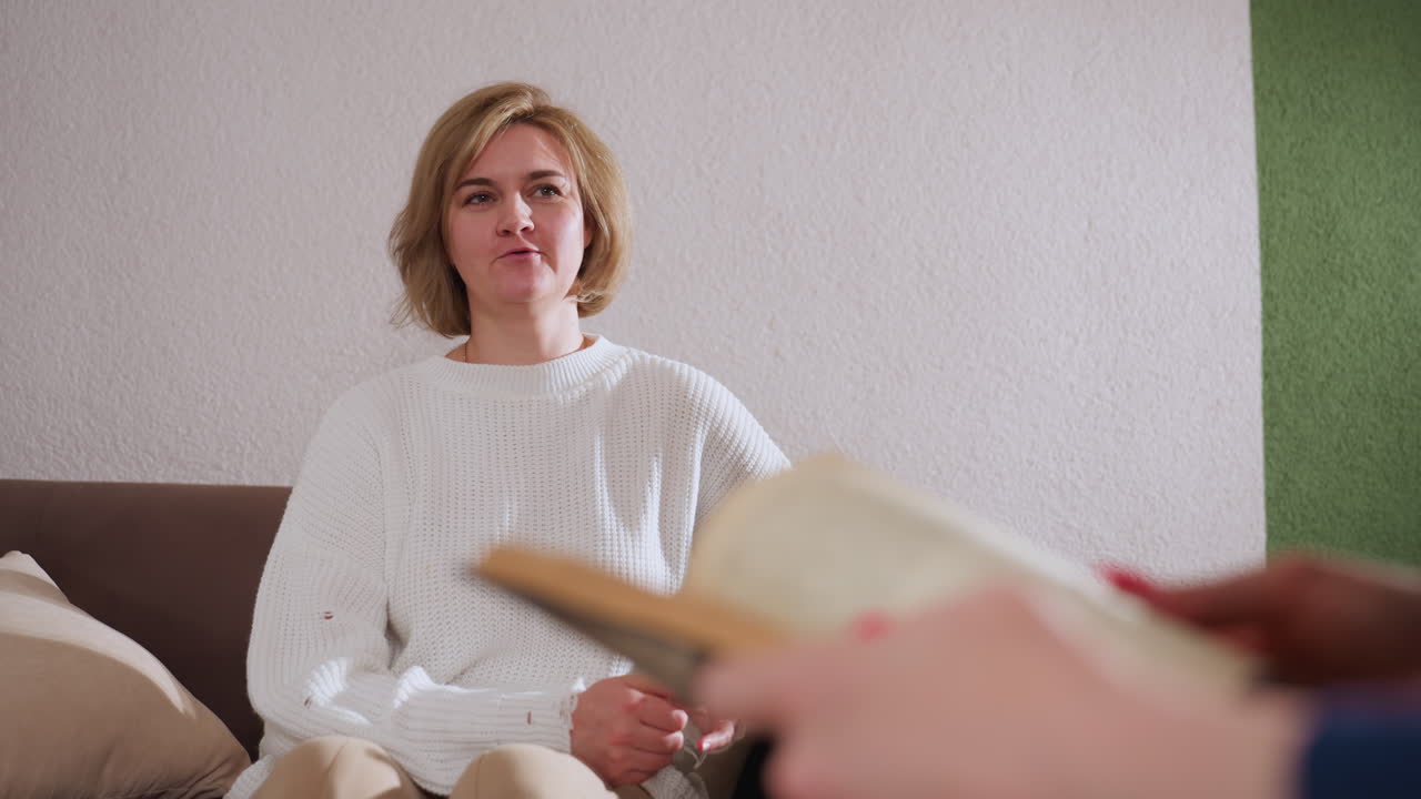 Cheerful client seated on sofa in white sweater expresses herself while engaging with clinical expert who flips through book pages during calm and supportive consultation in softly lit room