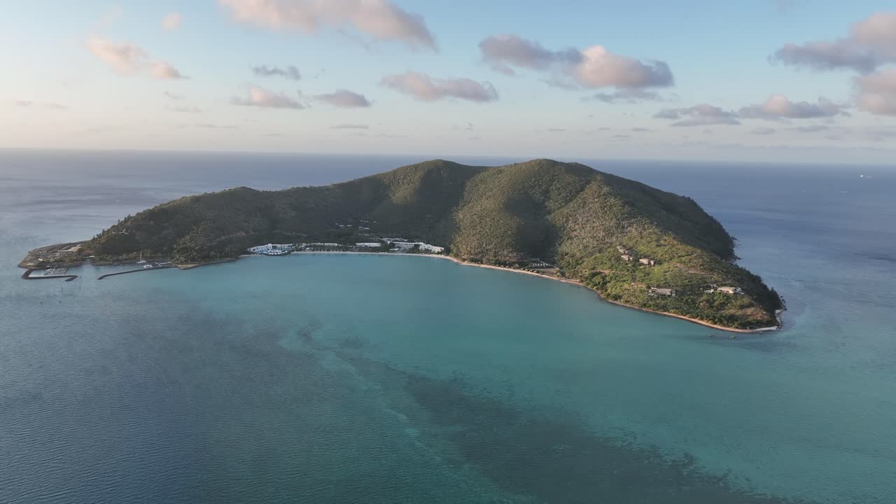 Ripresa lenta con drone all'indietro con una leggera elevazione all'ora d'oro, che si allarga su Hayman Island per rivelare la baia turchese, la costa del resort e le creste verdi illuminate dal sole, Whitsundays, Queensland, Australia