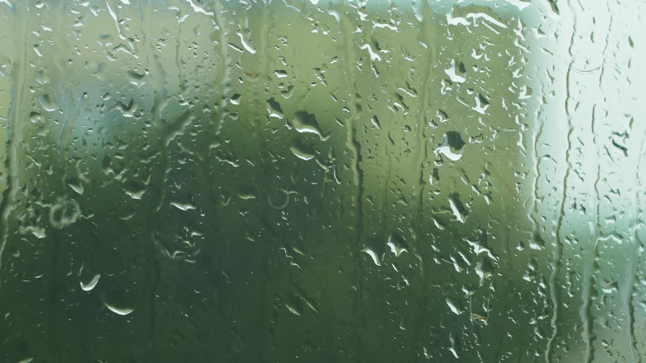 gotas de lluvia deslizándose lentamente sobre el cristal de la ventana en un día lluvioso, árbol desenfocado en el fondo, toma de primer plano medio