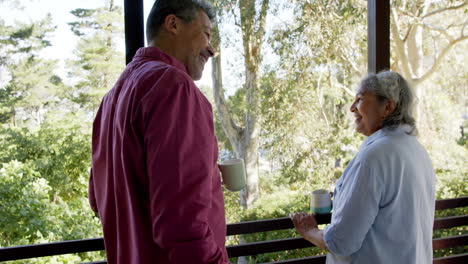 Happy-biracial-senior-couple-embracing-at-window,-talking-and-drinking-tea