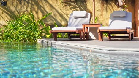 luxury lounge area for a two people rest with two deck chairs and umbrella next to the swimming pool surrounded with green plants background, low angle view from pool