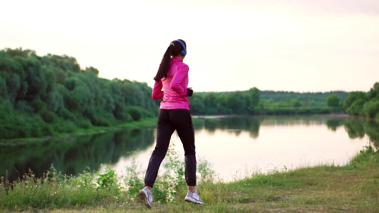 Brunette with long hair in headphones runs along the river in the Park in the morning at sunrise in the summer in a pink jacket and black pants