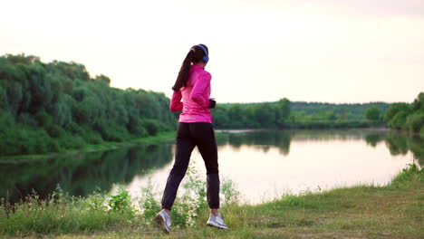 brunette with long hair in headphones runs along the river in the park in the morning at sunrise in the summer in a pink jacket and black pants