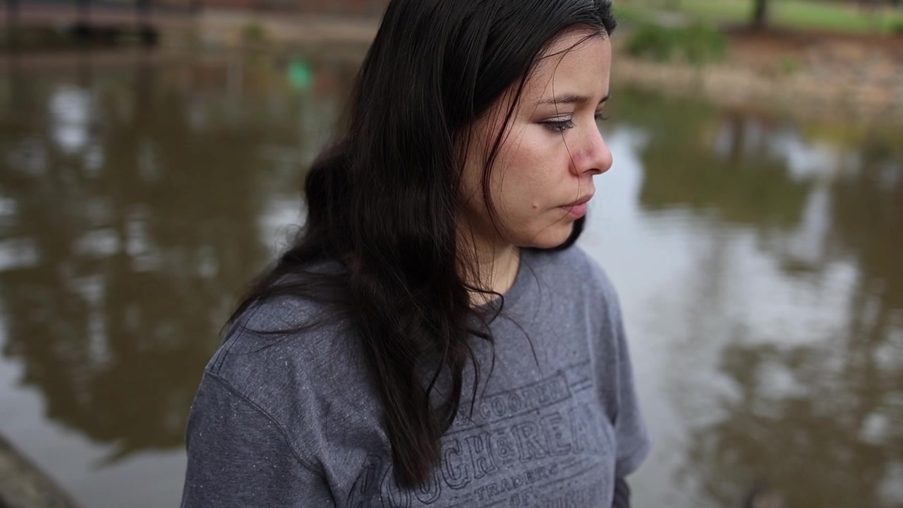 Uma jovem está pensativa ao lado de um lago tranquilo, perdida em pensamentos. Capturada em luz natural suave, este plano médio revela emoção e estado de espírito num cenário exterior exuberante.