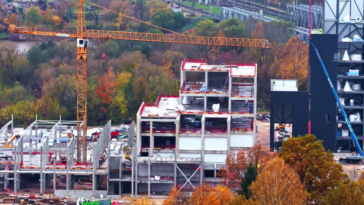 A Tall Crane Towers Over a Building Under Construction Surrounded by Autumn Trees - Aerial Pullback Shot