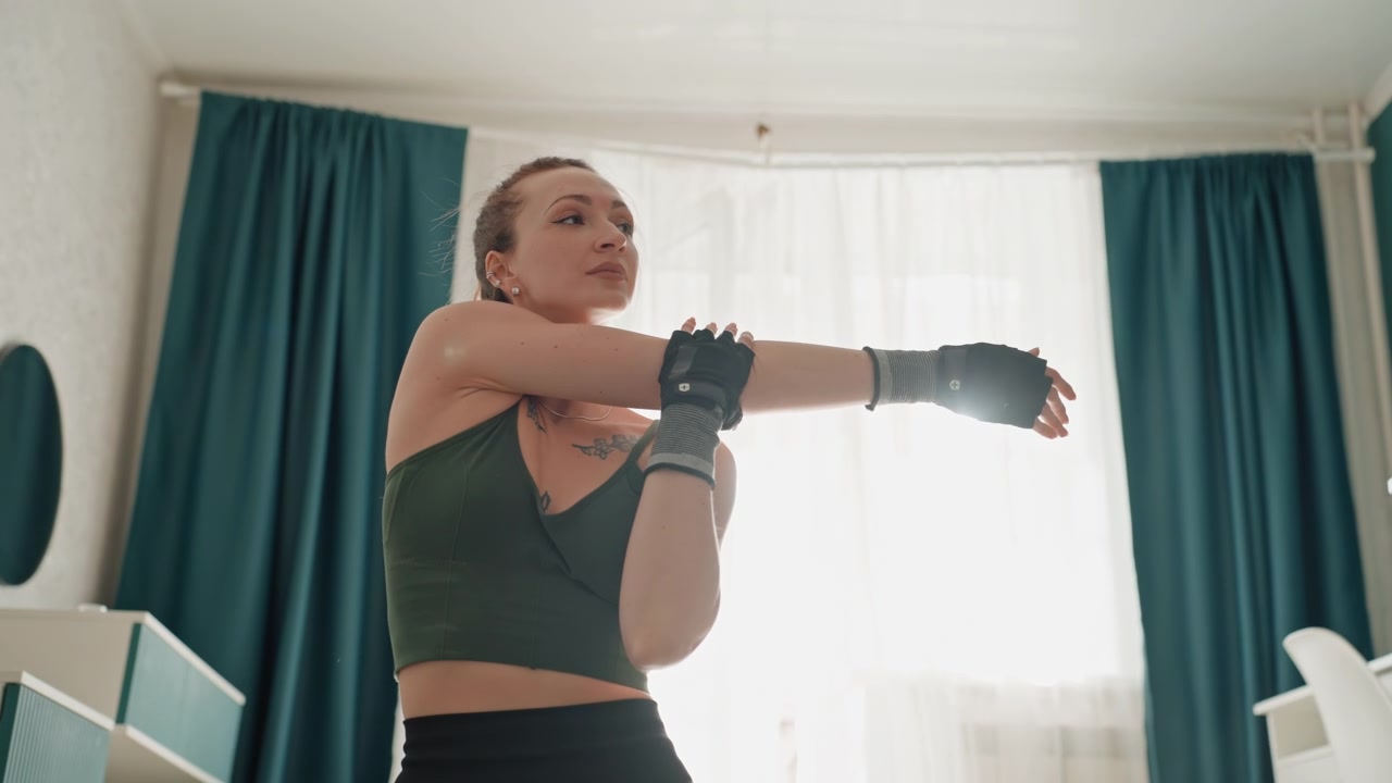 Jovem mulher enfaixando as mãos na sala de estar de casa, luvas sem dedos, cortinas iluminadas por trás pela luz solar, aquecimento focado antes do treino de boxe, alongamentos deliberados e proteção de pulso, atmosfera motivacional.
