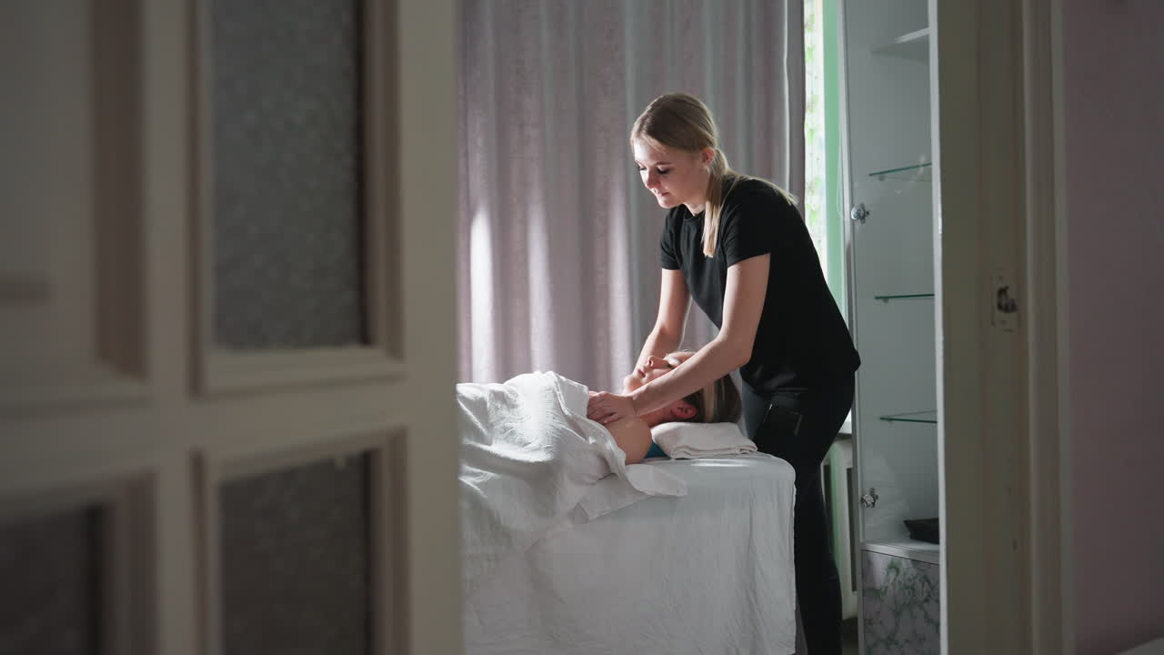 Massage therapist in black clothing giving chest massage to relaxed female client lying under white sheet in peaceful spa room with soft natural lighting, grey curtain, and glass cabinet in background