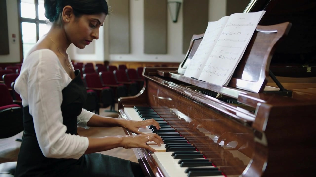 Camera pulling back showing pianist wearing layered outfit playing piano reading score at rehearsal