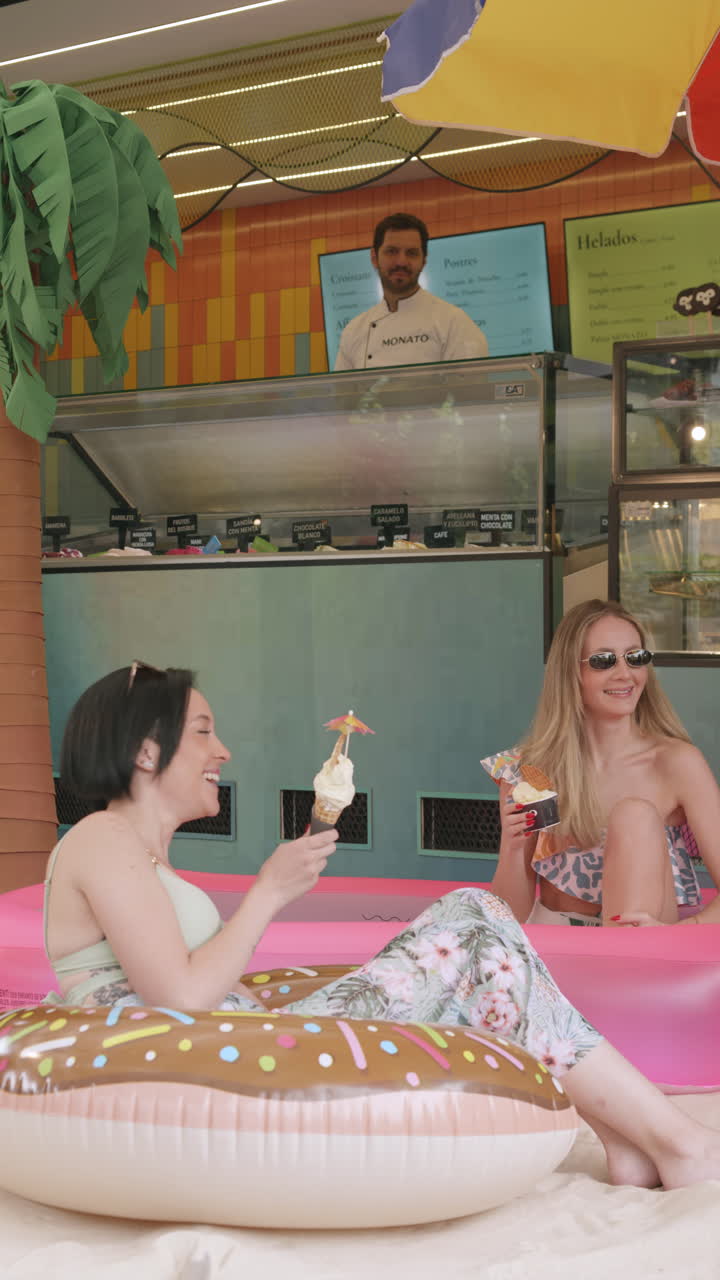 Two young women enjoy ice cream while relaxing in an inflatable pool in front of a modern ice cream parlor with tropical decor.