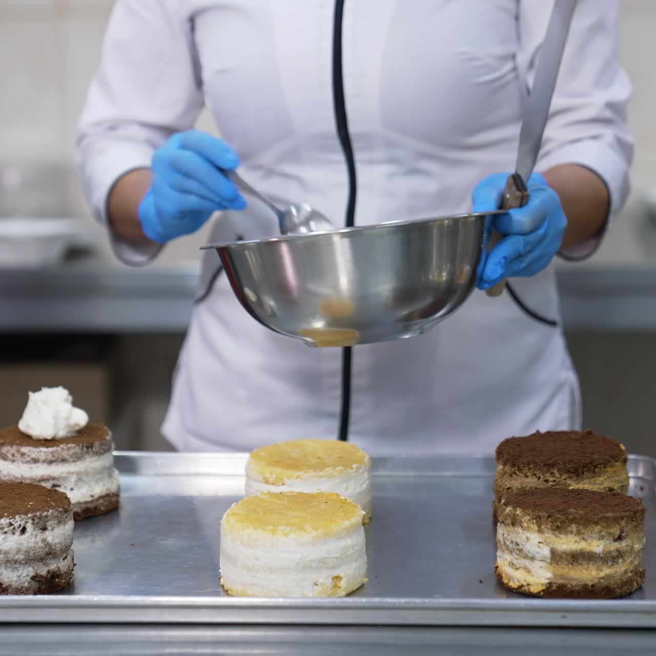 Little round cakes are put on the metal tray. Confectioner puts spoonfuls of cream on top of the cakes. Close up