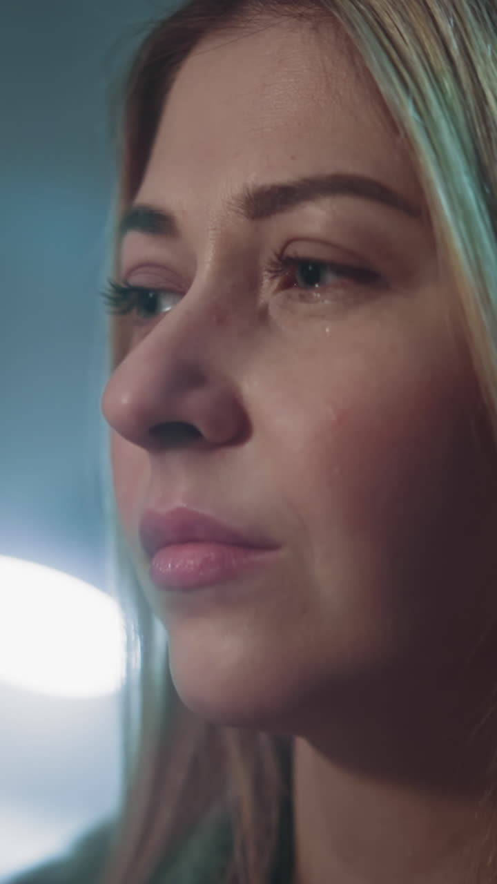 una mujer rubia preocupada y reflexiva sufre de una depresión severa en un fondo borroso en una habitación semi-oscura en casa, vista lateral extrema y cercana, cámara lenta.