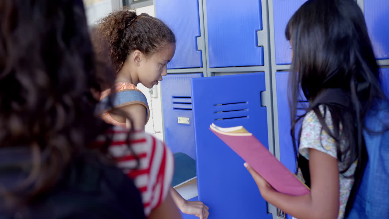 Three biracial girls are near blue lockers in a school, one holding a pink folder