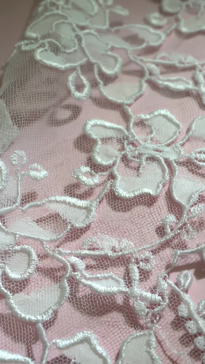 Vídeo vertical: câmera estabilizando e focando em renda bordada sobre fundo rosa, revelando o brilho da linha