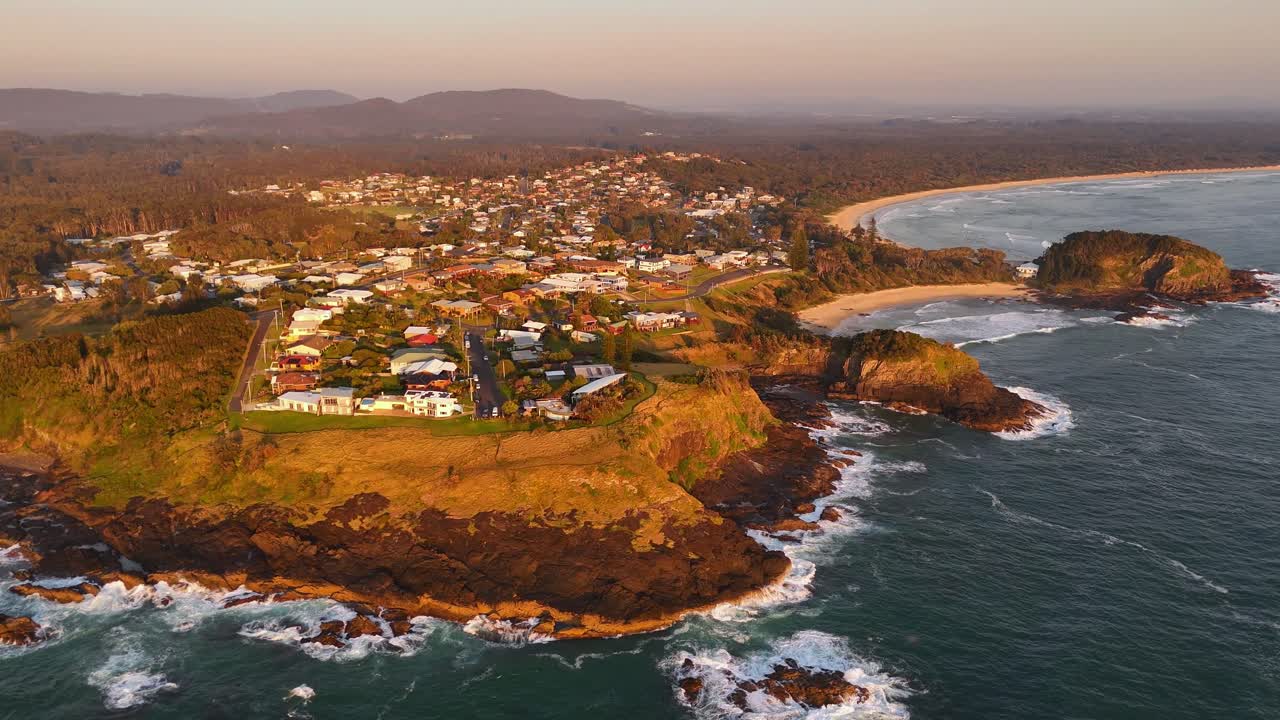 Drone aerial orbit of homes above beach and rocky headland at Scotts Head with foamy ocean surf at sunset