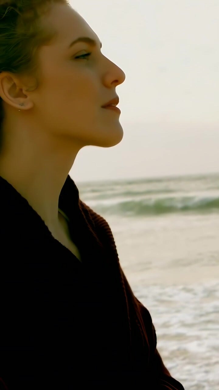 Vertical video: Standing woman in dark coat reflecting on shore as camera pulling back, copy space