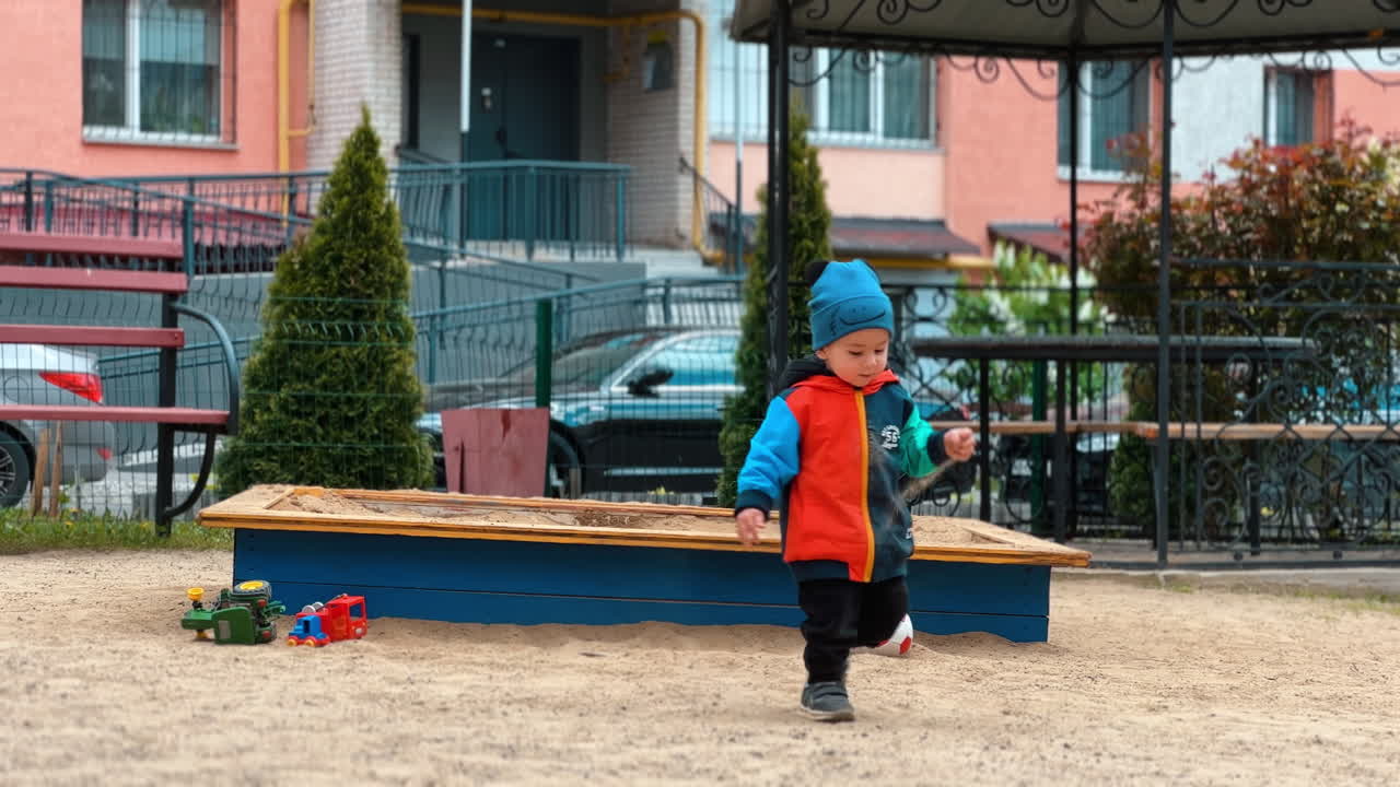 Baby boy in warm clothes bends to take some sand from the ground. Child runs by the playground throwing the sand.