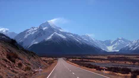 a long straight road leading to a large snow  mountain. the way to mount cook in new zealand. (driving pov)