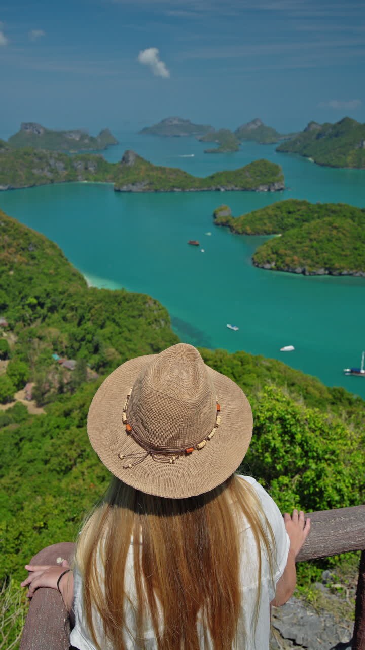 Joven con sombrero de paja de pie en un mirador de madera, mirando desde arriba el mar turquesa y las islas tropicales verdes. Vídeo en 4K de gran calidad.