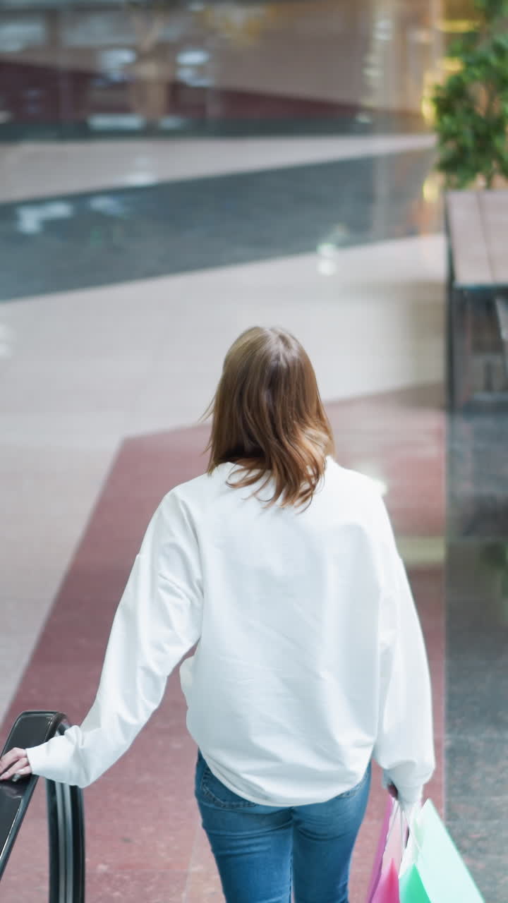 현대적인 건축물, 주변 환경을 반영하는 반이는 바닥, 우아한 디자인을 보여주는 넓고 밝은 조명 쇼핑몰에서 움직이는 에스컬레이터를 내려가는 여성의 뒷면