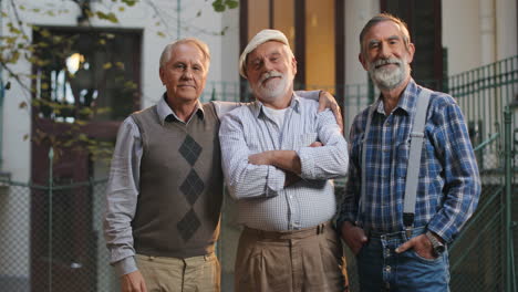 Foto-De-Retrato-De-Los-Tres-Amigos-Mayores-De-Pie-Y-Sonriendo-A-La-Cámara-Frente-A-La-Cámara-En-El-Patio