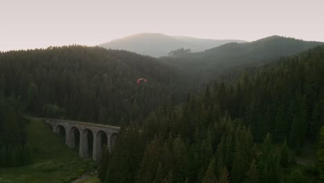 following a paraglider with a drone flying over a beautiful old bridge. chmarocsky viaduct in low tatras, slovakia, central europe. mountains in the background with a summer sunset.