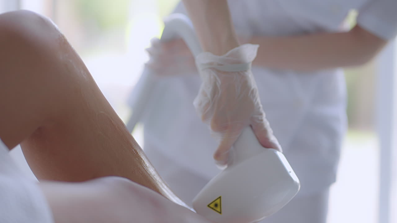 한 여성이 레이저 탈모 절차를 위해 소파에 누워 있습니다. 미용실에서 다리에 머리카락을 제거하는 하드웨어. 여성의 다리에 레이저 탈모를 제거합니다. 미용사의 손이 탈모를합니다.