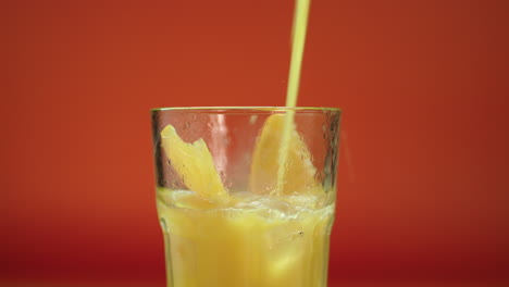 orange juice pouring in a highball glass filled with ice blocks and fresh oranges, isolated on an orange background