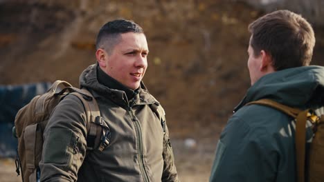 Close-up-over-the-shoulder-a-confident-man-with-a-short-haircut-in-a-dark-green-army-uniform-and-blue-medical-gloves-pats-his-military-friend-on-the-shoulder-during-their-communication-at-a-military-training-ground-with-large-black-tires-in-the-steppe