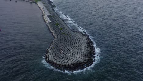 scenic top view of long stone rocky jetty in blue ocean sea water and waves crashing onshore, above circle aerial