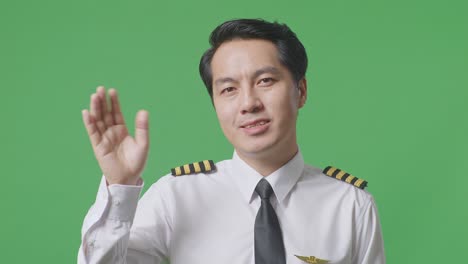 close up of asian man pilot smiling, waving hand, and saying bye while standing in the green screen background studio