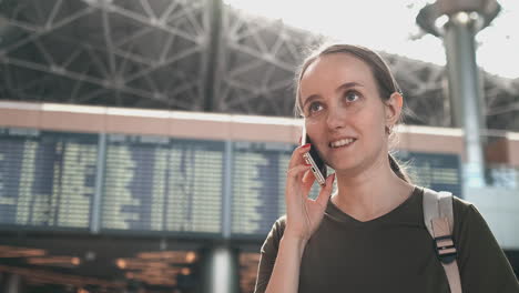 joven hermosa de pie en el aeropuerto llama al teléfono en el fondo del marcador con información sobre las salidas.